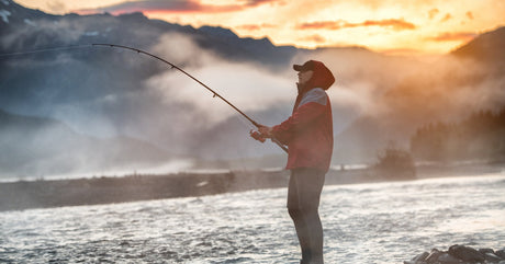 A man standing on a shoreline while fishing with the sun rising behind the mountains in the background.