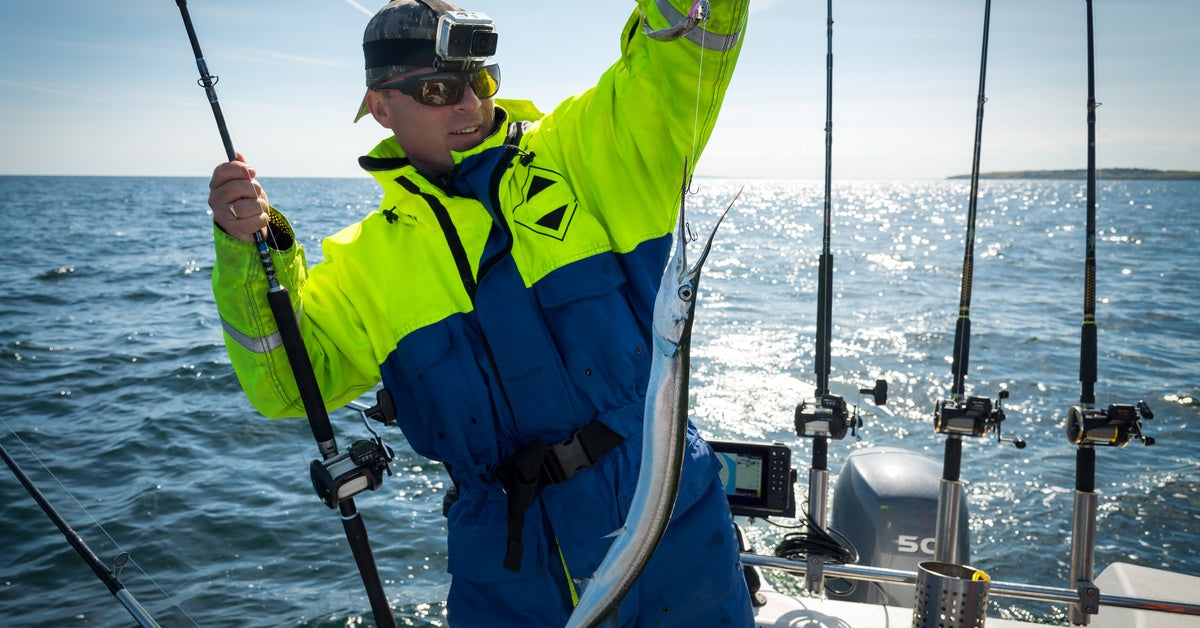 A man in a blue and hi-vis yellow coat holding up a fish he just caught while standing in a boat on open water.