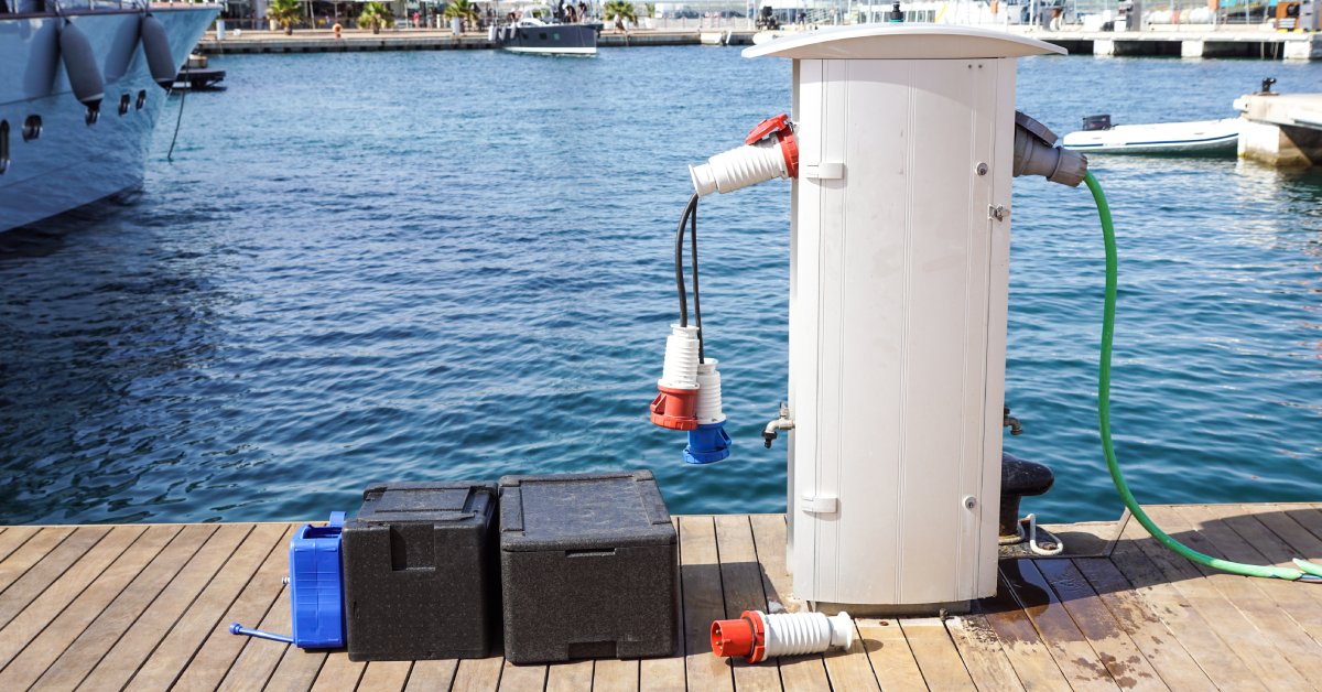 Two black marine batteries are sitting next to a power source on the dock of a marina. The water beyond the dock is dark blue.