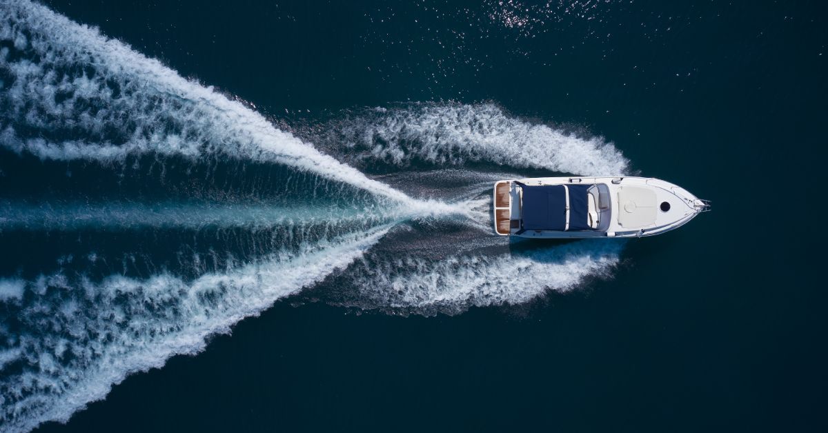 An overhead shot of a white boat moving through the water with lines of wake behind it. The sun glistens off the water.
