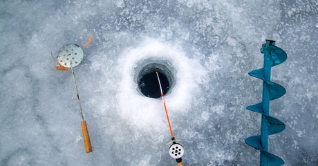 A hole in a thick sheet of ice with a fishing rod sitting in front of it next to a manual auger and another tool.