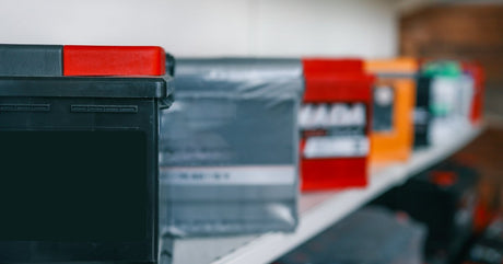 Various batteries are sitting on a white shelf. Some are wrapped in plastic, and one in the foreground is not.