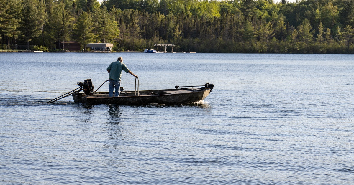 A man standing on his boat while using a trolling motor to navigate a lake with trees in the background.