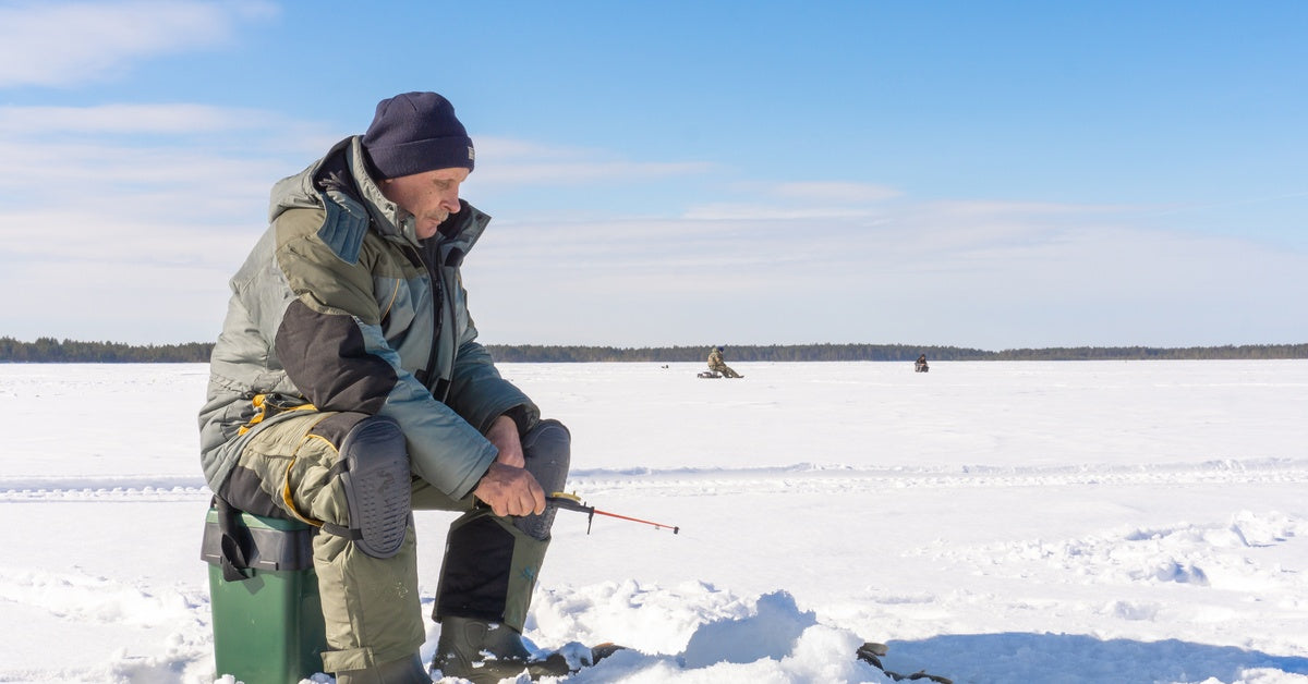 A man wearing a coat and black hat sitting on top of a green bucket on a frozen lake while ice fishing during the day.