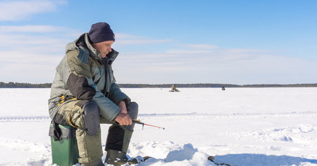 A man wearing a coat and black hat sitting on top of a green bucket on a frozen lake while ice fishing during the day.