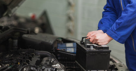 A mechanic wearing a blue jumpsuit standing in front of a car with the hood open while holding a battery.