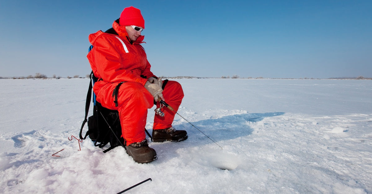 A man wearing insulated red clothing and sunglasses sitting on a chair with a pole while ice fishing during the day.