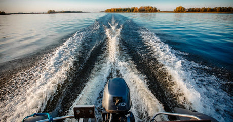 Waves form on a large body of water as a boat drives across the surface. Mature trees with fall-colored leaves line the shore.