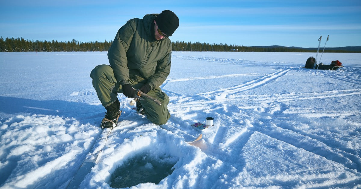 A male angler wearing an olive-green snowsuit, sunglasses, and a black beanie is holding a fishing pole above a hole in the ice.