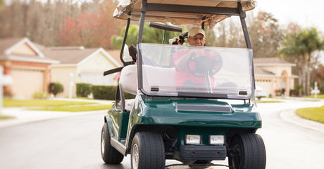An older man wearing a red shirt and tan baseball cap is driving a green golf cart down a residential street.