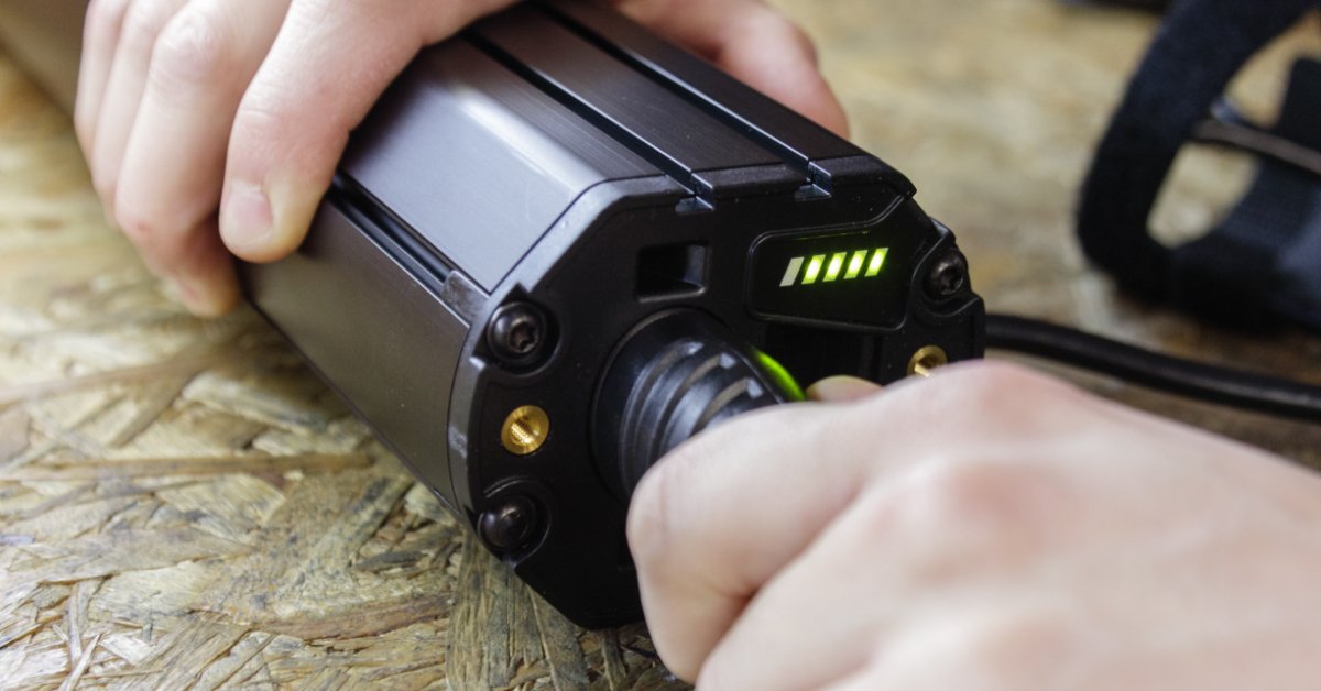 A close-up view shows a person plugging a black, cylinder lithium battery into a charging port. It sits on a wooden surface.