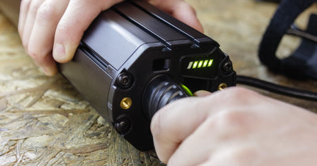 A close-up view shows a person plugging a black, cylinder lithium battery into a charging port. It sits on a wooden surface.