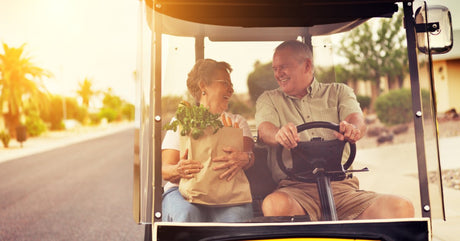 A smiling man is driving a yellow golf cart down the road. A smiling woman sits next to him holding a bag of groceries.
