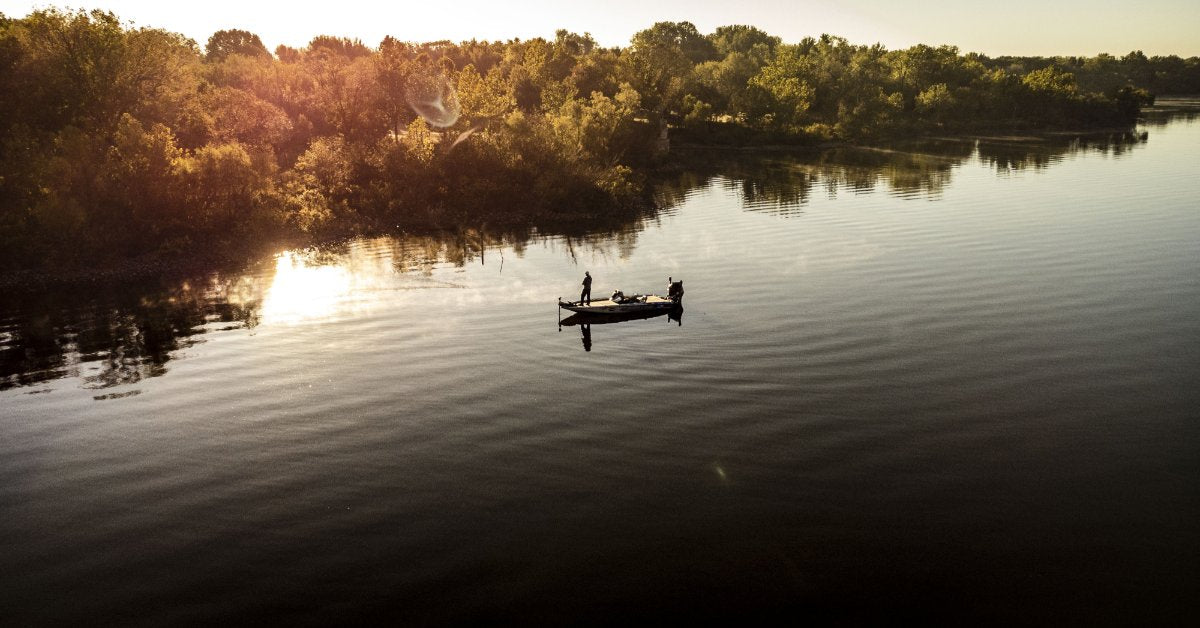 An aerial view shows a person standing up in a small fishing boat on a lake and holding a fishing pole.