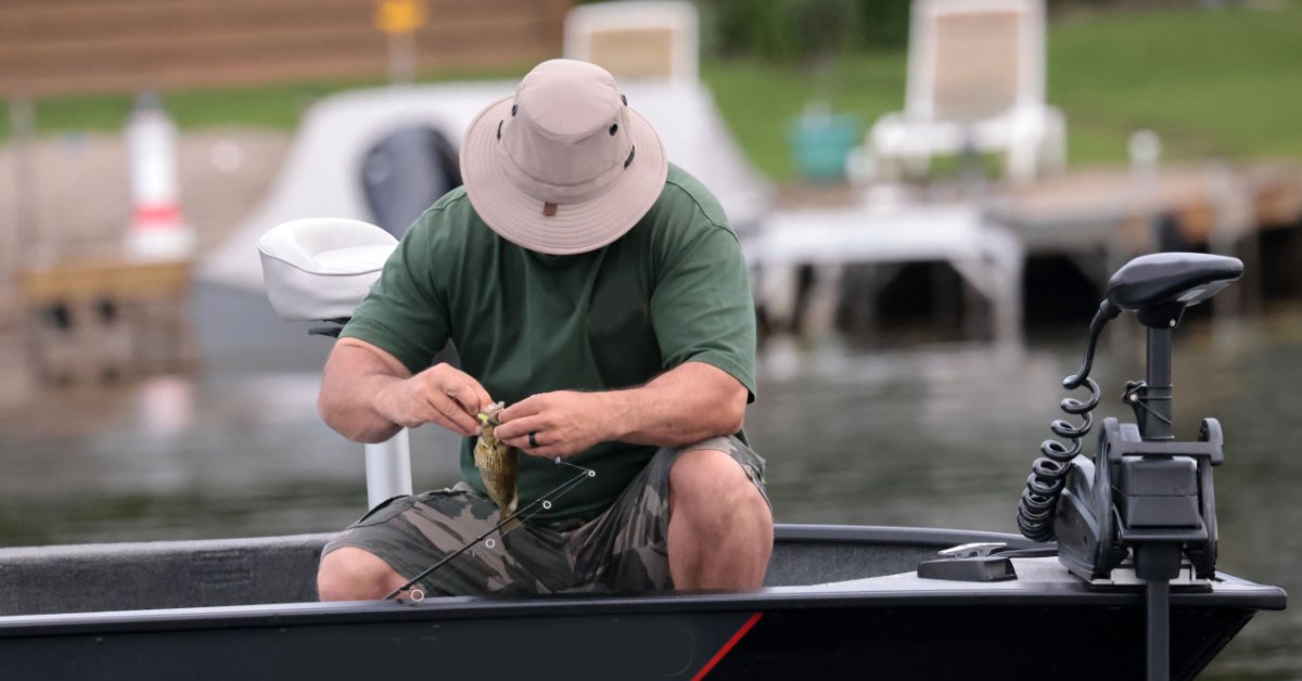 A male angler sits in a boat taking a small fish off a fishing hook. The boat has a black trolling motor.