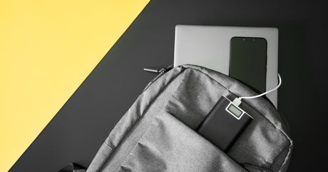 A gray backpack over a white and yellow background with an external battery in one of the pockets connected to a laptop.