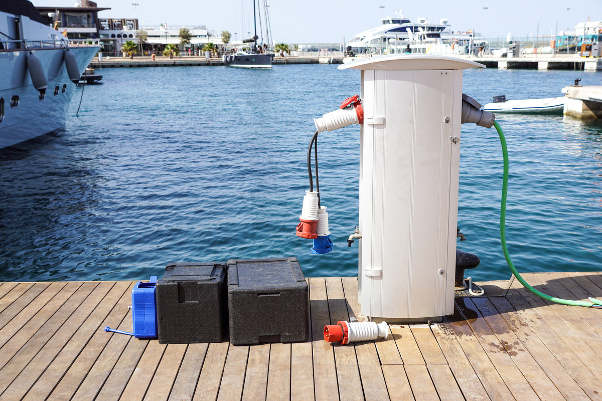 A boat charging station on a dock in a marina with a pair of marine batteries sitting next to it in plastic trays.