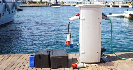 A pair of batteries in black casing sitting next to a white charger connected to cables on a dock in a marina.