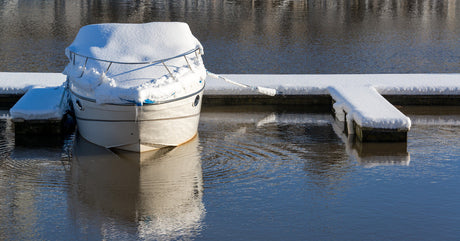 A boat tied to a dock on the water. It is covered with snow, and there are ripples bouncing off the keel.