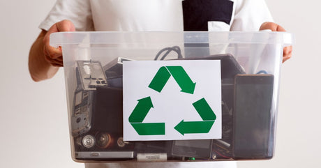 A person wearing a white shirt holding a tub of old electronics and batteries with a green recycling logo attached to it.