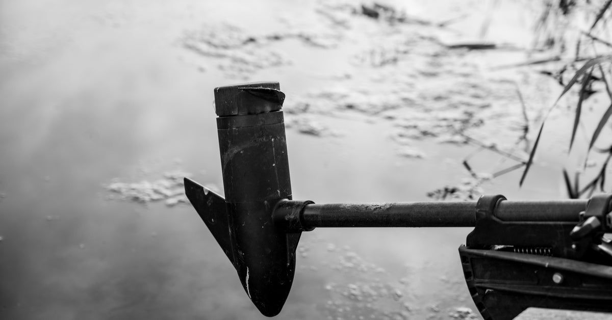 A black trolling motor pulled up out of a gray body of water on the back of a boat with moss and plants nearby.