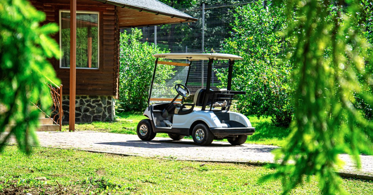 A golf cart parked in a gravel driveway in front of a wooden building surrounded by trees during the day.