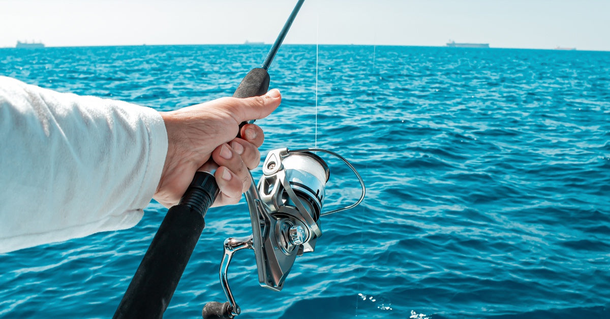 A person holding out a long black fishing rod by its handle, with the reel and line free above the blue ocean.
