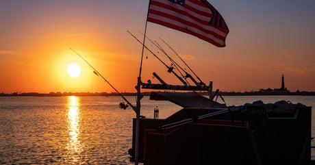 A boat with many rods hanging over its side sitting on the water at sunset. A pole with a US flag hangs over the boat.
