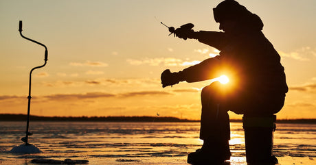 An angler sitting on a stool in front of a hole in a lake's ice sheet. They are lowering line into the cold water.
