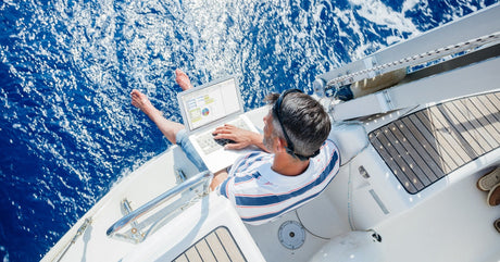 A person sitting on the edge of a sailboat with his feet over the water and using a laptop to review information online.