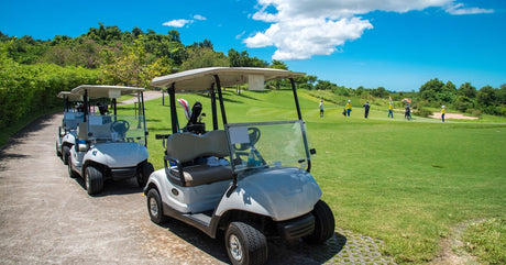 A row of white golf carts parked along the side of a green golf course with golfers standing on the green together.
