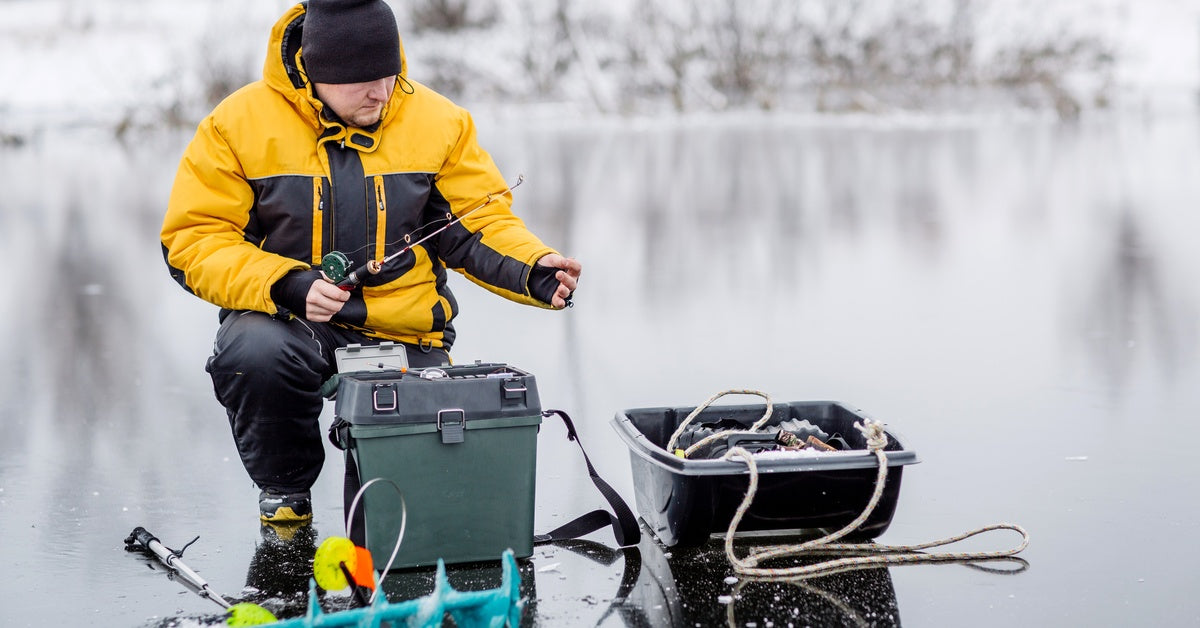 An angler in cold-weather attire sitting on the ice and organizing his setup while holding a small fishing rod in one hand.