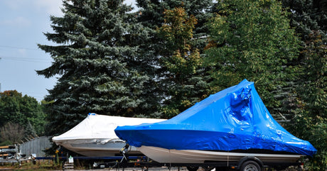 A pair of blue-and-white shrink-wrapped boats sitting in a parking lot with evergreen trees standing behind them.